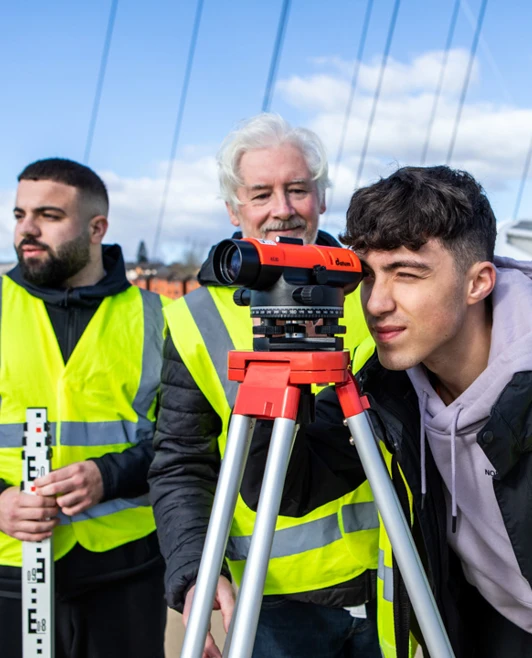 A young student in a high-visibility vest looking through a surveying instrument while the lecturer and another student observe and discuss measurements on a bridge A young student in a high-visibility vest looking through a surveying instrument while the lecturer and another student observe and discuss measurements on a bridge